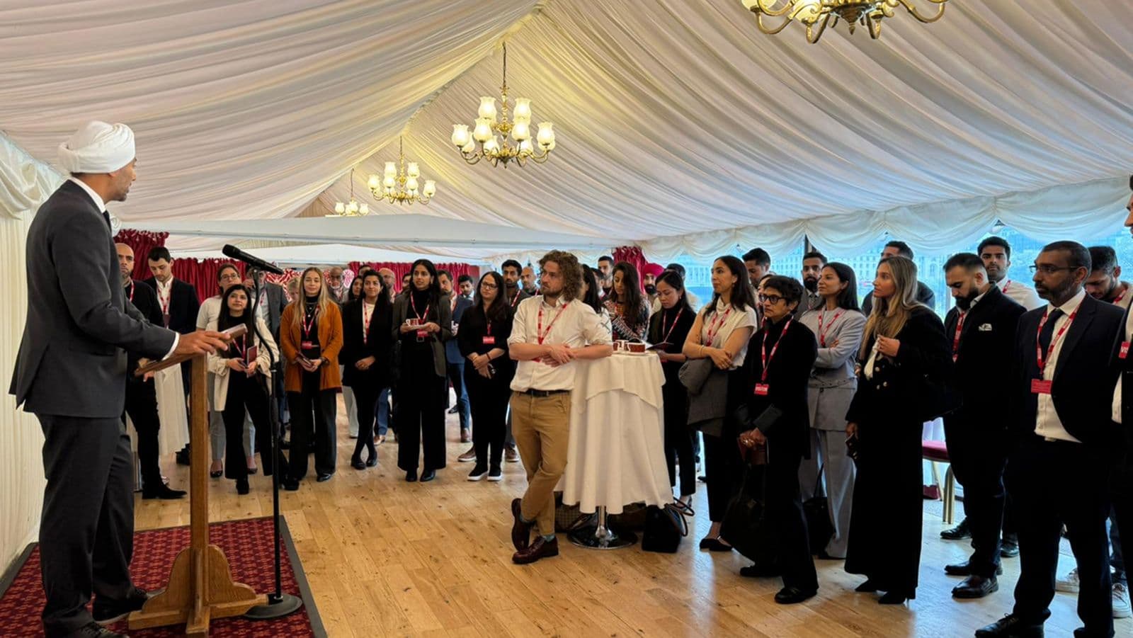 A speaker at a wooden lectern addressing a room of standing people, in a marquee with a draped ceiling and chandeliers.